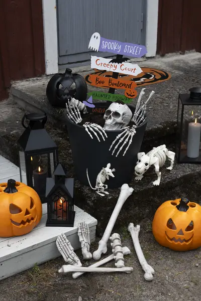 Luurankokoriste portailla halloween-koristeiden keskellä. Ympärillä on kurpitsoja, lyhtyjä, pääkalloja ja kyltti tekstillä "Spooky street".