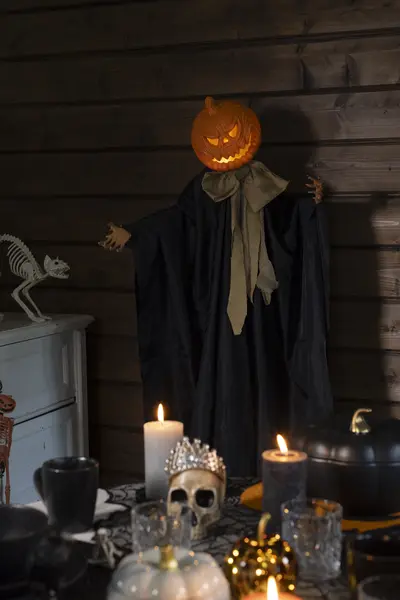Kurpitsapäinen hahmo osana näyttävää halloween-pöytäkattausta, jossa on kynttilöitä, pääkallokoristeita ja kurpitsoja.