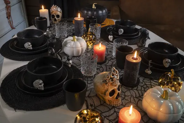 Näyttävä Halloween-pöytäkattaus, jossa on kynttilöitä, pääkallokoristeita, mustia astioita, koristekurpitsoita ja hämähäkinseittikangas.