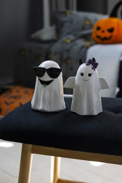 Kaksi valkoista haamukoristetta istuu tuolilla. Toisella haamulla on aurinkolasit, taustalla näkyy halloween-aiheisia koristeita ja kurpitsa.