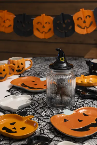 Halloween-juhlapöydän kattaus, jossa on kurpitsakuvioisia kertakäyttöastioita, koristeverkkoa sekä noitahattua kantava lasipurkkikoriste.