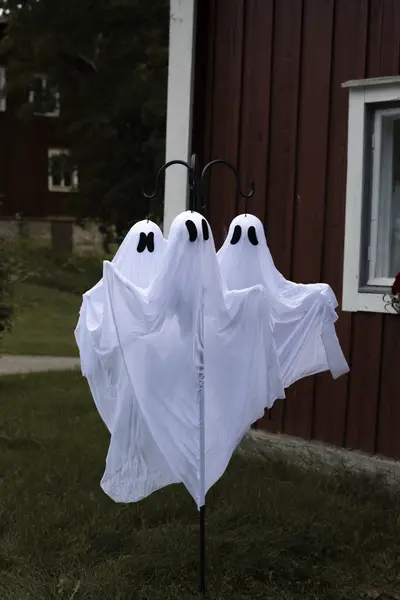 Kolme valkoista haamukoristetta puutarhatelineessä talon pihalla Halloween-sisustuksessa.