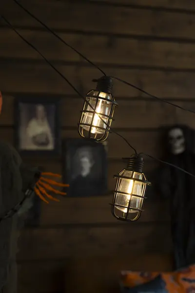 Lämpimästi hehkuvat riippuvat koristevalot Halloween-sisustuksessa. Taustalla näkyy kehystettyjä kuvia ja tummasävyisiä koristeita.