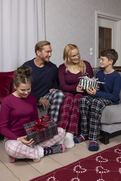 Perhe avaa lahjapaketteja olohuoneessa ruutukuosisissa pyjamahousuissa ja mukavissa kotiasuissa.